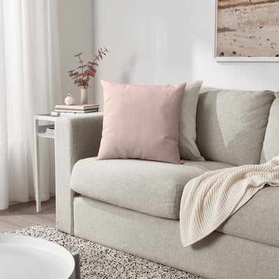 Cozy living room with gray sofa, pink/beige cushions, cream blanket, side table with vase and books.
