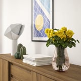 Glass vase with swirls holds yellow flowers on a sideboard, next to a lamp and cacti.