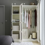Open white PAX wardrobe showcasing clothes, folded items, and storage boxes. Hangs centrally, fits neatly in room with dark grey wall.