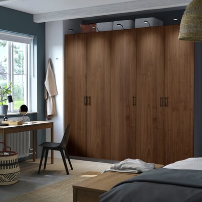 Large ikea PAX wardrobe in dark brown spans wall, includes doors, shelves, bins, with bed, desk, chair, and wicker basket.