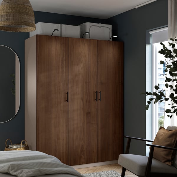 Modern bedroom with large wooden wardrobe. Dark walls, light bedding, cushioned seat. Wide window, plants.