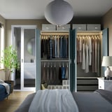 Well-organized bedroom closet with clothes, shelves, and a bed in front. Soft lighting and indoor plant.