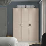 A beige PAX wardrobe in a bedroom. Its tall and wide, with four doors and mesh baskets on top, and stands beside a white bed.