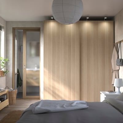 A modern bedroom with a PAX wardrobe in light wood, featuring sliding doors and embedded lighting. A grey bedspread and a cosy atmosphere are visible.