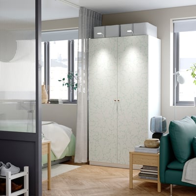 A modern, light-colored PAX wardrobe, larger than nearby furniture, with a floral pattern, two doors, and upper storage boxes.