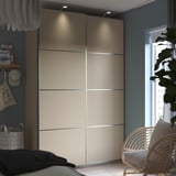 Large bedroom wardrobe PAX with cream sliding doors, underlit for modern feel. Next to a wicker chair and plant.