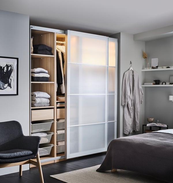 Modern bedroom with PAX wardrobe in white glass, grey bedding, black chair, and art.