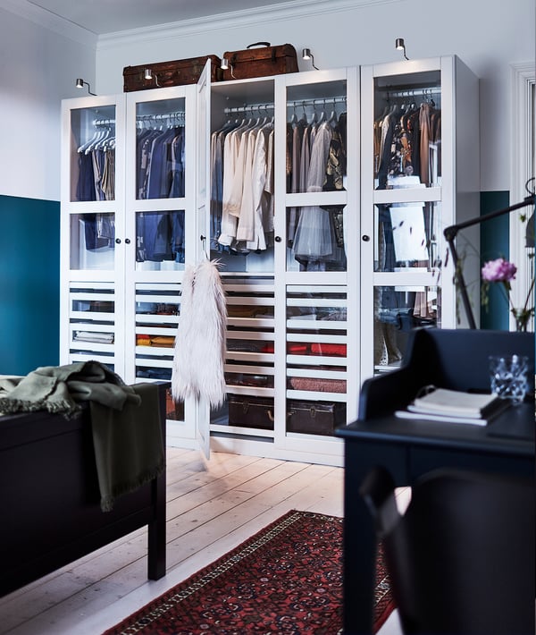 White PAX wardrobe with glass doors, clothes, and accessories. Wooden top, teal wall, patterned rug, organized drawers and shelves.