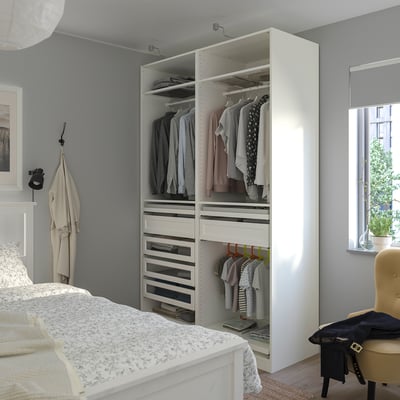 A PAX white wardrobe in a bedroom. Its tall, housing clothes and drawers, with a window nearby. A bed, chair, and coat hanger are visible.