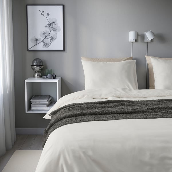 Grey bedroom with PILTANDVINGE duvet, white pillows, striped blanket. Bedside table with books, glass, photo, and wall lamp.
