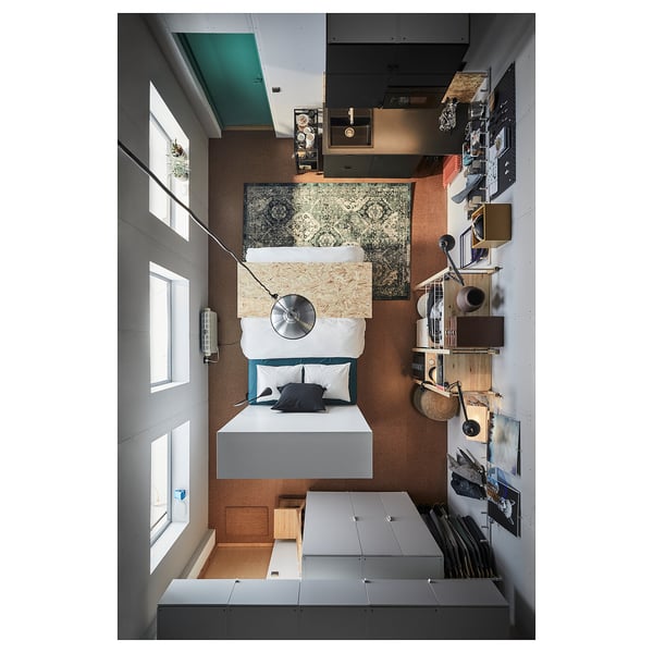 Modern minimalist bedroom: white PLATSA bed, gray bed frame, wooden shelves, hanging lights, patterned rug, large windows.