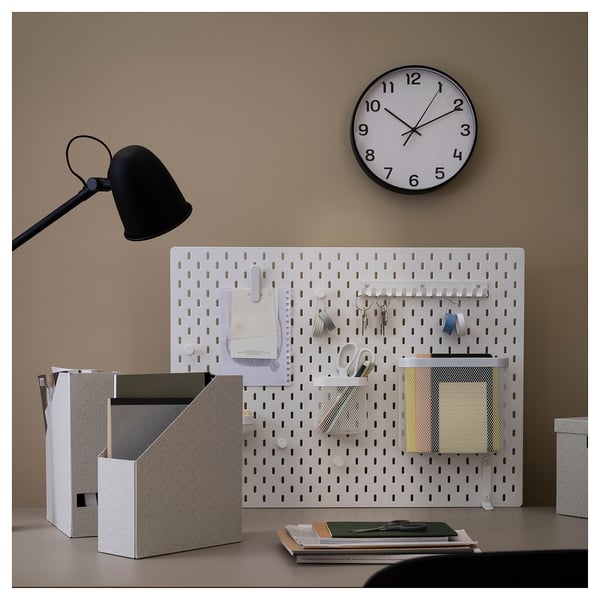 A minimalist office setup featuring a white PLUTTIS wall clock, a pegboard with storage, and a black desk lamp.