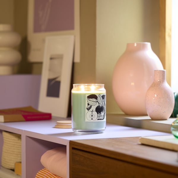 A green PRAKTHÄGER apple candle in a glass jar, surrounded by pink vases and books on a wooden shelf.