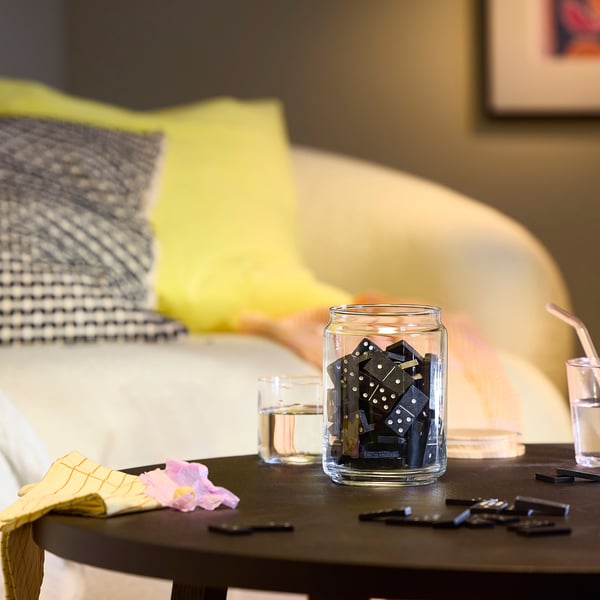 Clear jar of dominoes on wooden table near bed. Glass cup with straw and flower aside.