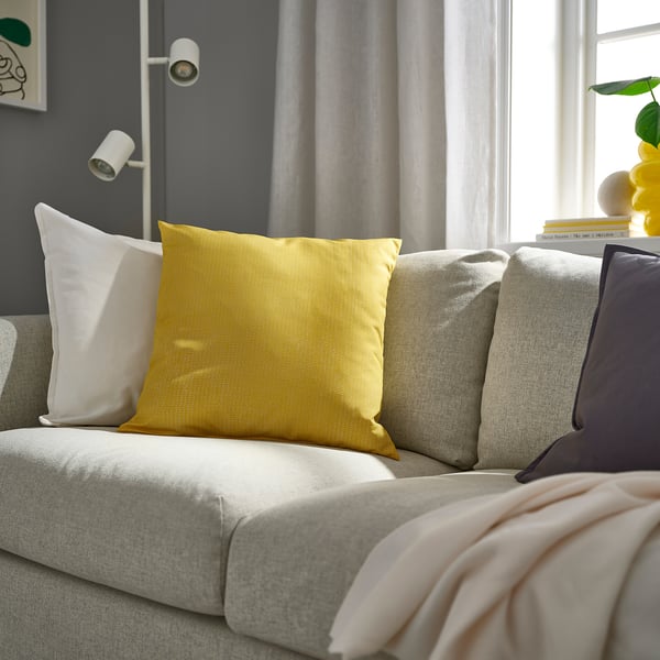 Modern living room with large yellow PROFETBLOMMA pillow, light grey sofa, white and purple pillows, pink throw, grey curtains, and wall sconce.