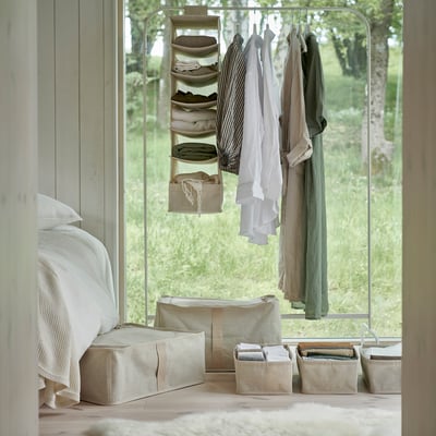 Bedroom with bed, window, and clothing rack. Beige organizers hold folded clothes; under-window storage for seasons.
