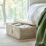 Beige fabric storage box by bed, stores clothes, green blanket nearby, protects from dust.