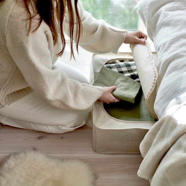 A person opening a beige storage case on a wooden floor, ideal for dust-free storage of seasonal items, with handles and ventilation holes.