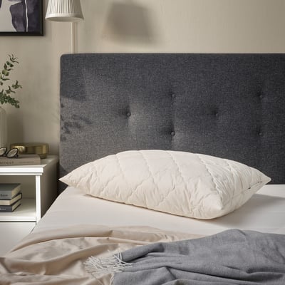 RÅDHUSVIN pillow on bed, white, quilted, between book & dark grey headboard.