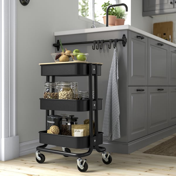 Small black RÅSKOG trolley with 3 shelves, fitting under counters, holding jars and fruits, next to gray cabinets.