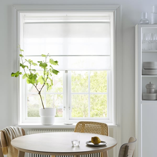 Modern dining area with white RINGBLOMMA blinds, a wooden table, and wicker chair. A plant sits by the window and a lemon on the table.