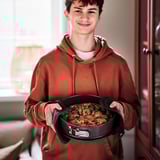 Teenager in brown hoodie holds black pan with baked food, likely removed from oven using oven mitts.