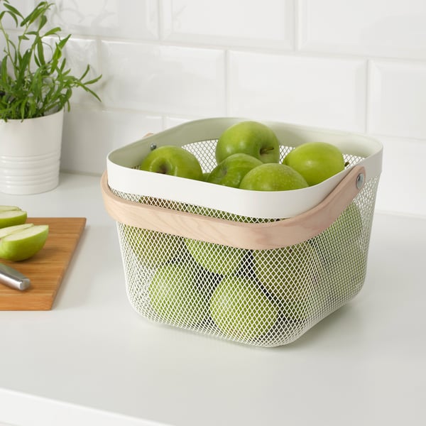 A mesh storage basket with wooden handles holding green apples. It ventilates to keep produce fresh. On a white countertop near a cutting board.