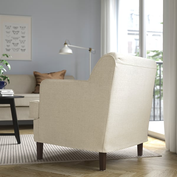 Beige armchair with wooden legs in cozy room; lamps, sofa, and art visible.