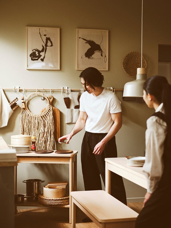 Two people cook in a modern kitchen. A man organizes dishes by a trolley, a woman arranges plates at a table.