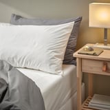 Crisp white bedding set with wooden nightstand. Sheets and pillows in neat arrangement. Compact lamp, book, pen on table. Bedding fits thick mattress.