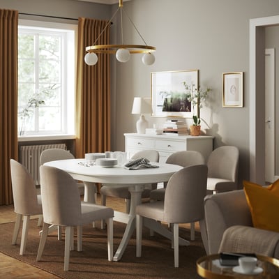 Modern dining space with white ROSENTORP chairs in beige around a round table, under a circular chandelier. Linen curtains and a sideboard with decor.