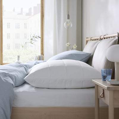 A cozy, well-lit bedroom a bed with a white ROSENVIAL pillow protector, blue pillows, and a wooden bedside table with a lamp and glass.