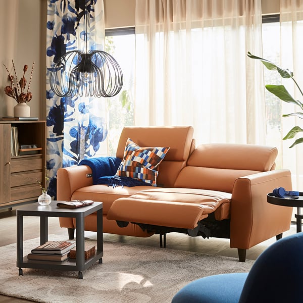 Orange leather recliner sofa near window with floral curtains, blue blankets, multi-colored pillows, and a small coffee table with books and a vase.