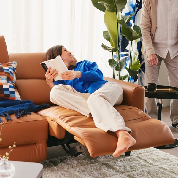 Person relaxing in RULLERUM recliner, reading a book while adjusting backrest. Brown leather, comfortable and supportive, ideal for lazy evenings.