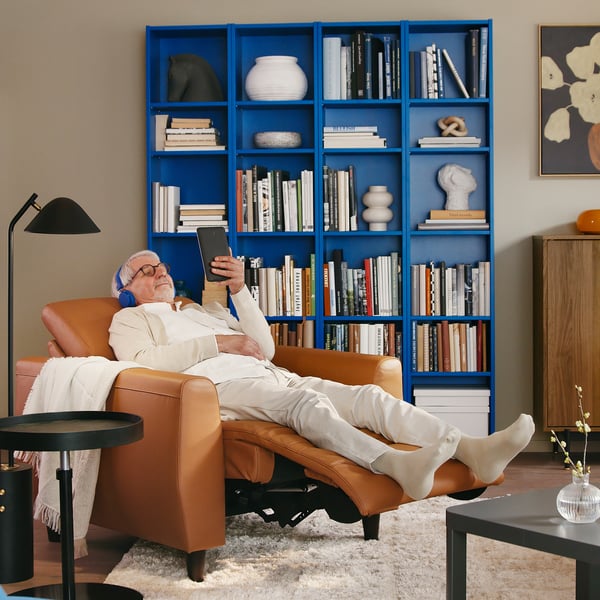 Person reclining in brown leather RULLERUM electric recliner, reading on tablet, surrounded by books and decor.