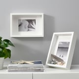 Two SANNAHED frames: one on wall, one on shelf with books, holding flower and beach photos; plant beside books.