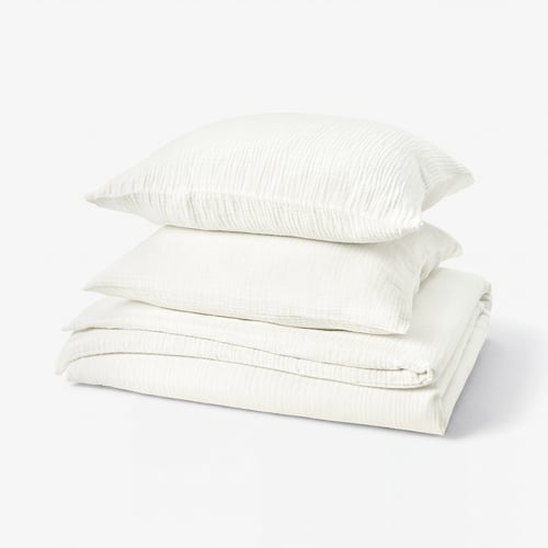 Stacked white bedding set, including folded duvet and pillows, featuring soft, textured muslin fabric.