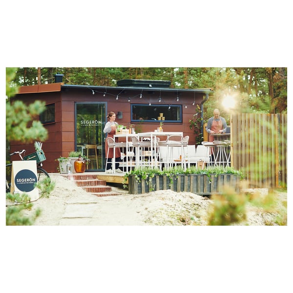 Small outdoor café, brown wood paneling, white tables and chairs. Woman serving, man seated. Lush green background, sandy ground.