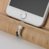 A smartphone rests on a wooden holder, which has a cable hole for charging.
