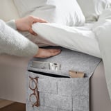 A person in a gray sweater sits on a bed beside a gray felt storage pocket with a checkered pattern and mesh squares, holding a white pillow.