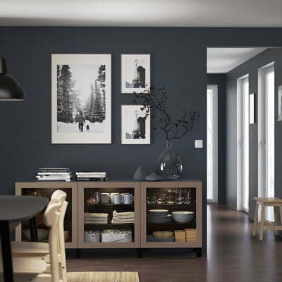 Modern dining room with SINDVIK cabinet, glass doors, black pendant light, framed art, wooden chair, and vase with branches.