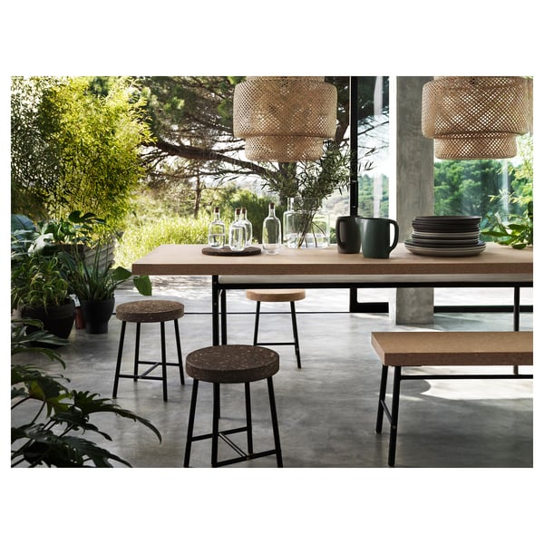 Modern natural dining setting with bamboo pendant lights, cork table, and black stools surrounded by greenery.