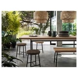 Modern natural dining setting with bamboo pendant lights, cork table, and black stools surrounded by greenery.