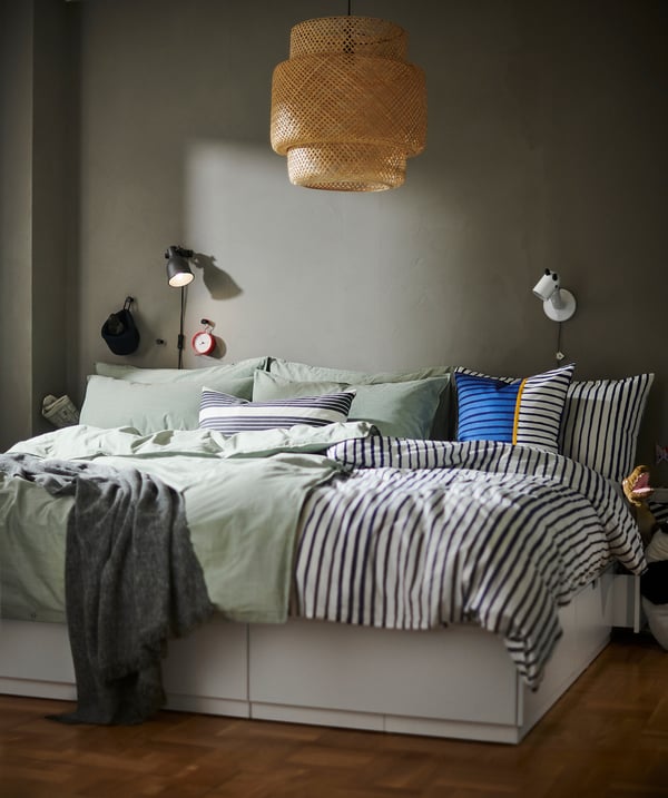 Cozy bedroom with SINNERLIG bamboo lattice lampshade casting warm light, striped bedding on white bed, grey walls, and wooden floor.