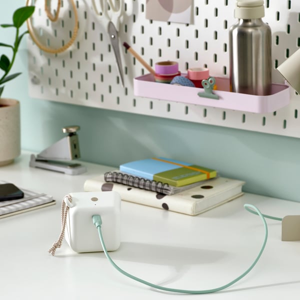 A white desk with a green usb-c cable connected to a small device, notebooks, and a plant. A pegboard with office supplies in the background.