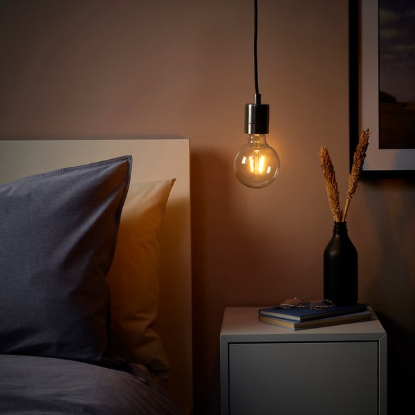 A cozy bedroom with a SKAFTET pendant lamp, black vase with dried plants, books, and a blue pillow.