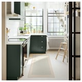 Modern kitchen with dark green cabinets, white tiles, and a striped beige and blue runner. Large windows allow natural light.