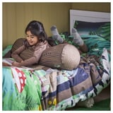 Little girl reads on bed with SKOGSDUVA support pillow. Tree-trunk shaped cushion aids comfort.