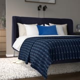 Modern bedroom with large bed. Dark blue headboard & checkered blanket. White rug, white slippers. Nice & cozy setting.