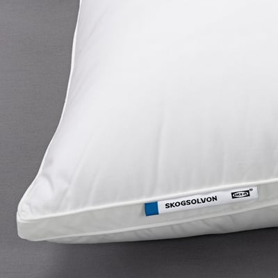 Close-up of a SKOGSOLVON pillow on a bed. White, plush, with visible brand tag.